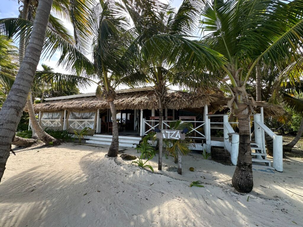 Open-air dining area at Itza Resort on Lighthouse Reef Atoll Belize surrounded by palm trees and white sand near the water’s edge